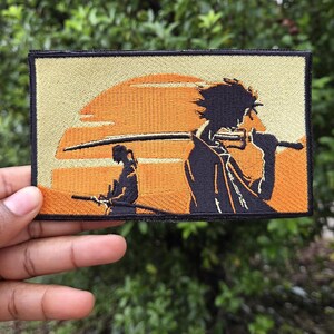 May include: Embroidered patch featuring a stylized anime scene with two figures and a sunset backdrop. The patch has a black border, with orange and tan colors dominating the design. The figures are silhouetted, holding swords.