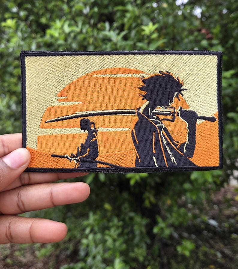 May include: Embroidered patch featuring a stylized anime scene with two figures and a sunset backdrop. The patch has a black border, with orange and tan colors dominating the design. The figures are silhouetted, holding swords.