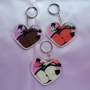 May include: Heart-shaped keychains with a cartoon illustration of a butt, wings, and horns. The keychains have a pink border and a silver keyring. The butt illustration comes in brown, red, and white.