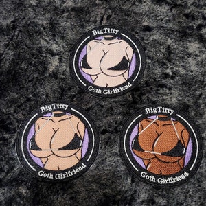 Peut inclure: Trois écussons ronds brodés avec une bordure noire. Chaque écusson présente une illustration d'un buste en haut de bikini noir, avec le texte "Big Titty" au-dessus et "Goth Girlfriend" en dessous. Le buste est illustré en trois tons de peau différents.