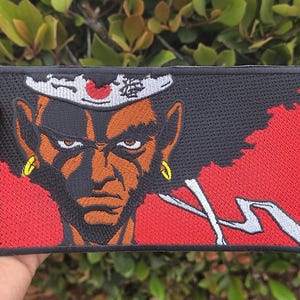 Op de afbeelding: Geborduurde patch met een gestileerde illustratie van een vastberaden personage met een afro. De patch heeft een zwarte achtergrond met rode en witte accenten. Het personage heeft een hoofdband met Japanse tekst.