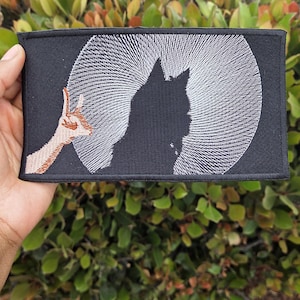 May include: Black rectangular patch featuring a silhouette of a cat against a white radial design, resembling a moon. A brown and tan hand gesture is on the left. The patch is held against a backdrop of green foliage.