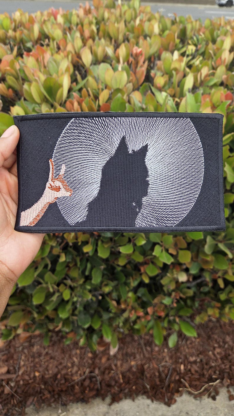 May include: Black rectangular patch featuring a silhouette of a cat against a white radial design, resembling a moon. A brown and tan hand gesture is on the left. The patch is held against a backdrop of green foliage.