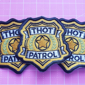 May include: Embroidered patches in a shield shape with the words "THOT PATROL" in white lettering. The patches have a gold star and laurel wreath design, with a navy blue border and gold accents. The patches are on a pink grid background.