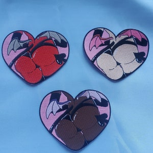 Può includere: Tre toppe ricamate a forma di cuore. Ogni toppa presenta un design a cartone animato di un sedere con ali di pipistrello e un'imbracatura. Le toppe sono nei colori rosso, beige e marrone, con accenti rosa e neri.