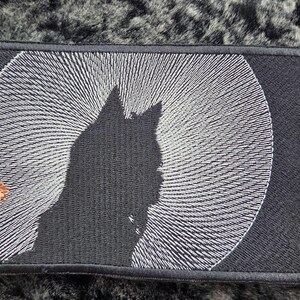 May include: Black rectangular patch featuring a cat silhouette against a white spiral design. A hand with a peace sign is on the left. The patch is next to a coin, all on a dark, textured surface.