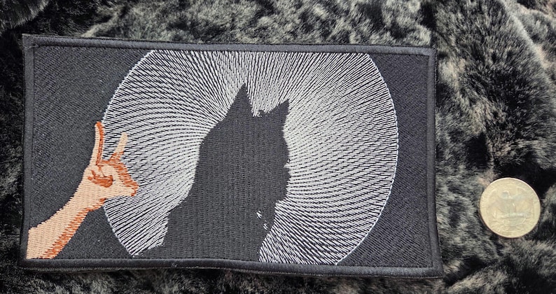 May include: Black rectangular patch featuring a cat silhouette against a white spiral design. A hand with a peace sign is on the left. The patch is next to a coin, all on a dark, textured surface.