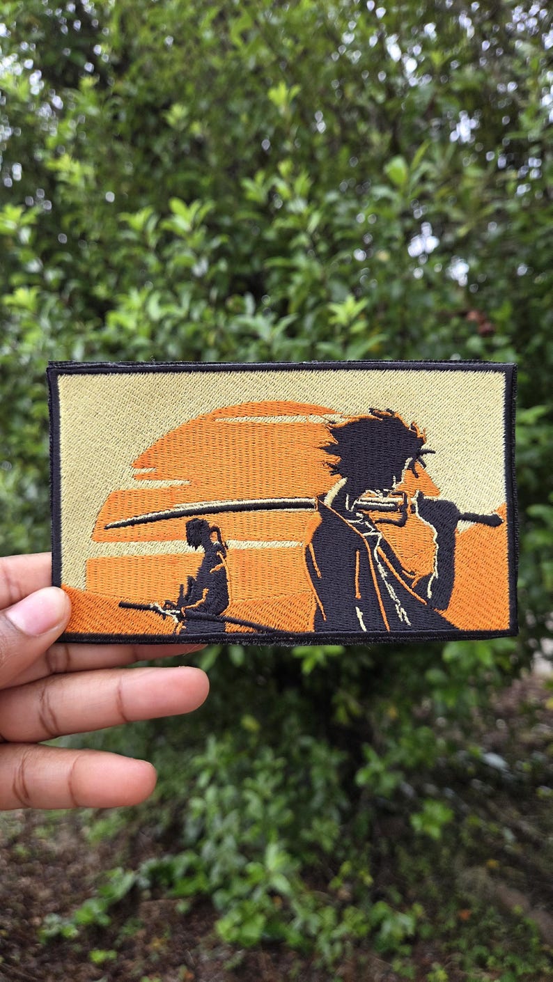 May include: Embroidered patch featuring a stylized scene with two figures and a sunset. The patch has a rectangular shape with a tan border and a background of orange and tan. The figures are in black, holding swords.