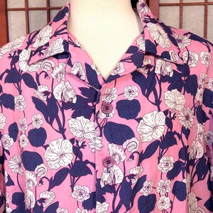 May include: Pink button-down shirt with a floral pattern. The shirt features white flowers and dark blue leaves and stems against a pink background. The shirt has a classic collar and matching pink buttons.