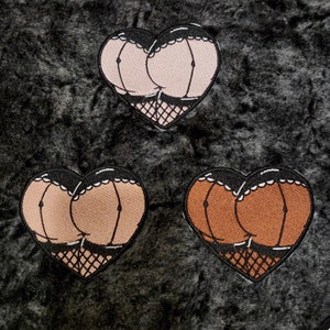May include: Three heart-shaped embroidered patches with a stylized rear view design. Each patch has a black border with a white scalloped edge. The buttocks are in shades of tan and brown, with black lines and fishnet stockings.