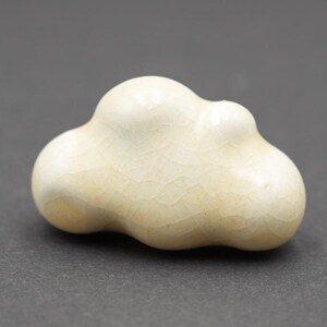 May include: A small, off-white ceramic object shaped like a stylized cloud with a smooth, glossy finish and a visible fine crackle glaze pattern throughout its surface. The piece features rounded, organic contours and a soft, cream-colored tone.