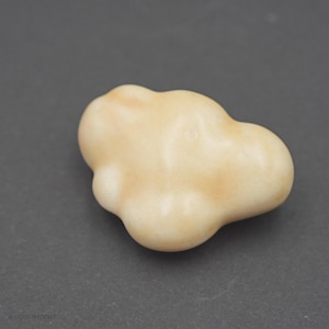 Könnte beinhalten: Ein kleines, glattes, cremefarbenes Objekt mit einer unregelmäßigen, abgerundeten, wolkenartigen Form. Die Oberfläche weist subtile Farbvariationen von hellem Beige bis Cremeweiß auf und hat ein mattes Finish. Der Text A CLOUD IN POCKET ist sichtbar.
