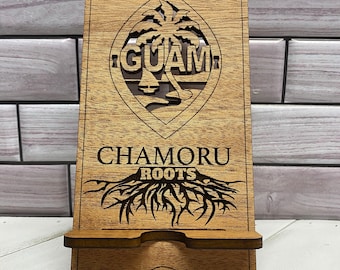 May include: Wooden phone stand with a cutout of the Guam seal, featuring a palm tree and the word "GUAM". Below the seal is the text "Chamoru Roots" with a stylized tree root design.