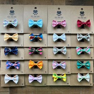 May include: A display of twenty colorful bow ties for pets, each attached to a cardboard backing. The bow ties feature various patterns, including polka dots, floral prints, and geometric designs. The cardboard backing has the text "PETS AND LOVE" and a paw print logo.