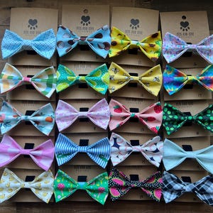 May include: A selection of patterned bow ties, featuring designs such as gingham, fruit, and floral prints. Each bow tie is displayed on a brown card with a paw print and heart logo. The bow ties are arranged on a wooden surface.