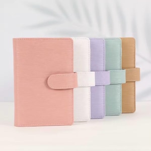 May include: A row of pastel-colored, textured leather notebooks with matching snap closures. The notebooks are in shades of pink, white, lavender, mint green, and tan. The notebooks are arranged side by side.