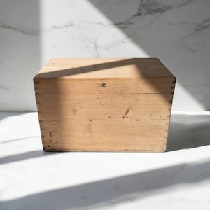 Puede incluir: Una caja rectangular de madera clara con vetas visibles y esquinas ensambladas con juntas de dedos. La caja cuenta con una tapa plana y un pequeño cierre metálico circular centrado en el panel frontal de madera de tono natural.