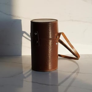 Op de afbeelding: Een cilindrische bruine leren etui met een handvat. De etui is afgesloten met een deksel en heeft een textuur. Het handvat is een lichtere bruintint. De etui staat rechtop op een reflecterend oppervlak.