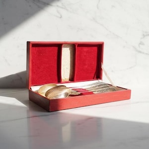 Puede incluir: Una caja roja con interior de terciopelo contiene cubiertos antiguos. El juego incluye cucharas y un tenedor, con mangos ornamentados. La caja está abierta, revelando el contenido sobre un fondo de mármol blanco.