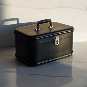 Puede incluir: Un neceser rectangular negro con ribete y asa plateados. El estuche tiene un cierre plateado y está sobre una superficie de mármol. El estuche se refleja en la superficie.