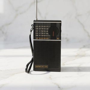 Op de afbeelding: Een zwarte vintage radio met een zilveren antenne die omhoog is uitgeschoven. De radio heeft een draaiknop en een luidsprekerrooster. De tekst "НЕЙВА РП-205" is zichtbaar op de voorkant. Een zwarte riem is aan de zijkant bevestigd.