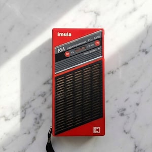 Op de afbeelding: Een vintage rode en zwarte draagbare radio met de merknaam "imula" bovenaan. De radio heeft een zwart luidsprekerrooster en een zwarte draagriem. De voorkant van de radio heeft de tekst "AM" en andere Cyrillische tekst.