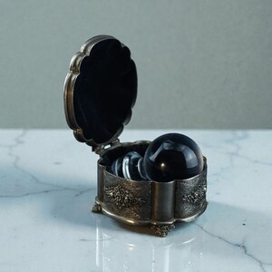 May include: An open, silver-coloured jewellery box with a black velvet lining. It contains a large, black crystal ball and two clear glass spheres. The box has floral detailing and is on small feet. The box is made of metal.
