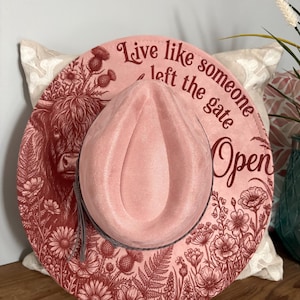 May include: Pink cowboy hat with a floral and highland cow design. The hat features the text "Live like someone left the gate open". The hat has a gray band and tassel.