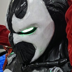May include: A full-body costume featuring a black and white mask with glowing green eyes, a textured black chest piece with a large white emblem, a silver skull-shaped shoulder accent with green light, and a flowing red cape draped over the shoulders.