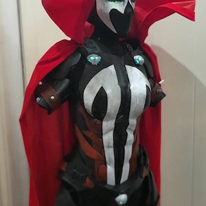 May include: A detailed Spawn costume with a black and white mask, glowing green eyes, and a red cape. The costume features a black and brown armored design with white accents and a chest emblem.