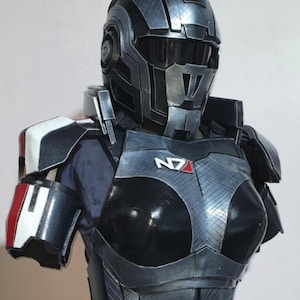 Peut inclure: Une armure de cosplay noire et grise détaillée, comprenant un casque et un plastron. L'armure présente le logo "N7" et des accents rouges et blancs sur l'épaule. Le design est futuriste et angulaire, avec un aspect métallique.