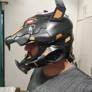 以下が含まれることがあります： 犬のようなデザインの黒と金のコスプレヘルメット。ヘルメットは、オレンジ色のアクセントと赤く光る目を持つ、詳細で質感のある表面が特徴です。デザインは未来的で様式化されています。