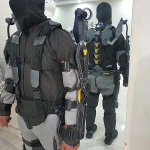 Puede incluir: Un traje t&aacute;ctico futurista negro y gris con detalles amarillos. El traje incluye una cobertura facial completa, un torso de malla y varios paneles protectores. El dise&ntilde;o incorpora un sistema de arn&eacute;s y componentes articulados.
