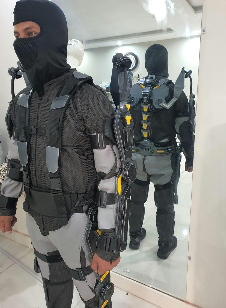 Puede incluir: Un traje t&aacute;ctico futurista negro y gris con detalles amarillos. El traje incluye una cobertura facial completa, un torso de malla y varios paneles protectores. El dise&ntilde;o incorpora un sistema de arn&eacute;s y componentes articulados.
