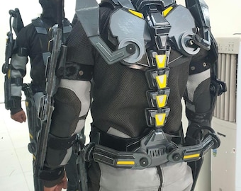May include: A futuristic exoskeleton suit in shades of gray, black, and yellow. The suit features a complex design with articulated joints, a spine-like structure, and protective plating. The person wearing the suit is also wearing a black balaclava.