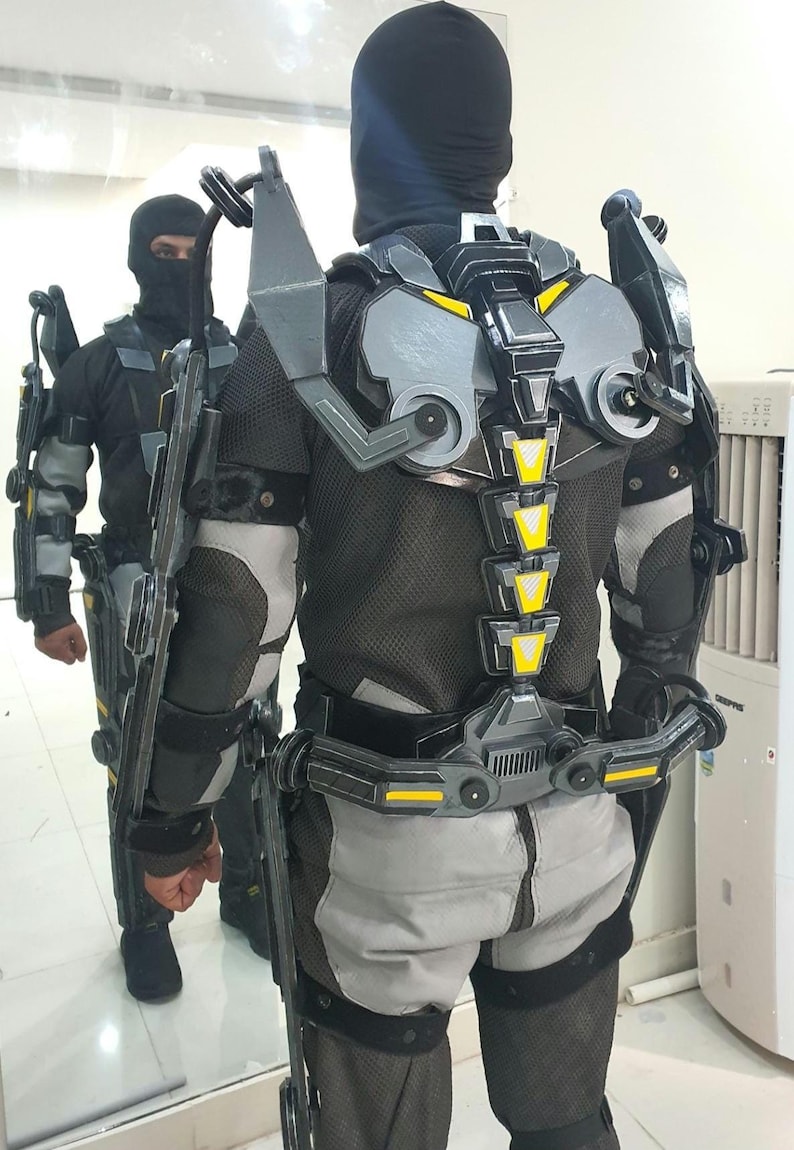 Puede incluir: Un exoesqueleto futurista en tonos grises, negros y amarillos. El traje presenta un dise&ntilde;o complejo con articulaciones, una estructura similar a la columna vertebral y placas protectoras. La persona tambi&eacute;n lleva un pasamonta&ntilde;as negro.