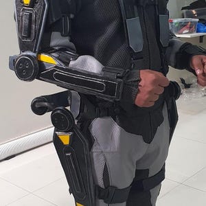 Puede incluir: Un traje de exoesqueleto negro y gris con detalles amarillos. El traje cubre los brazos y las piernas, con articulaciones y correas. La persona lleva un pasamonta&ntilde;as negro y una chaqueta de malla negra.