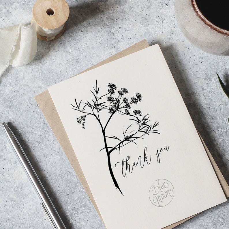 May include: A white card with a black line drawing of a flower and the text "thank you" written in cursive. The card also has a small circle with the text "Blue Moon" in cursive.