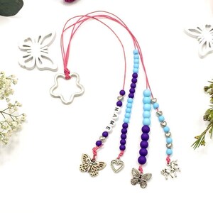 Może przedstawiać: Zakładka do książki z koralikami w kolorze różowym, fioletowym i niebieskim, z zawieszką w kształcie srebrnej gwiazdki, motyla, serca i jednorożca. Koraliki tworzą napis "NAME".