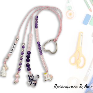 Może przedstawiać: Brelok z różowymi i fioletowymi koralikami z zawieszką w kształcie serca i różnymi ozdobami, w tym kotem, jednorożcem i królikiem. Brelok jest oznaczony jako "NAME".