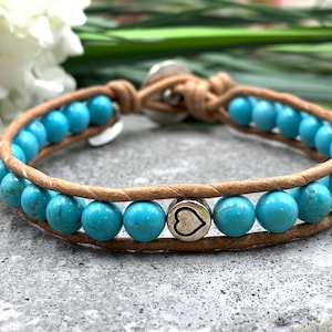 Könnte beinhalten: Ein türkisfarbenes Lederarmband mit einem Herz-Anhänger. Das Armband besteht aus türkisfarbenen Perlen und braunem Lederband. Der Herz-Anhänger ist silberfarben und hat ein Herz-Design.