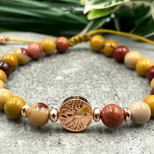 Könnte beinhalten: Ein Armband aus natürlichen Steinperlen in Braun-, Gelb- und Rosatönen. Die Perlen sind auf einer gelben Schnur aufgefädelt und haben einen roségoldenen Lebensbaum-Anhänger.