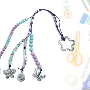 Może przedstawiać: Naszyjnik z koralików w kolorze fioletowym i niebieskim z zawieszką w kształcie gwiazdy i różnymi srebrnymi zawieszkami, w tym motylami i kwiatami. Naszyjnik ma wyraz "NAME" napisany białymi koralikami.