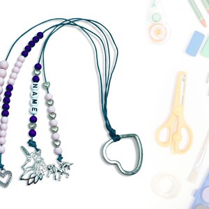 Może przedstawiać: Trzy koraliki naszyjniki z zawieszkami. Naszyjniki są wykonane z różowych i fioletowych koralików i mają srebrne zawieszki w kształcie serc, jednorożców i imienia napisanego białymi koralikami. Naszyjniki są przymocowane do turkusowego sznurka ze srebrnym zapięciem w kształcie serca.