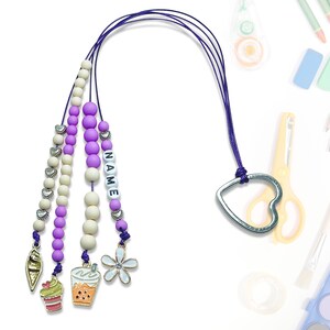 Może przedstawiać: Sznur z koralików w kolorze fioletowym i białym z srebrnym zawieszką w kształcie serca i etykietą z imieniem "NAME". Sznur ma trzy zawieszki: babeczkę, bubble tea i kwiat.