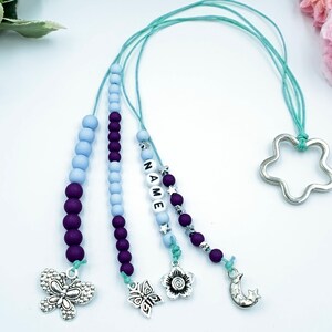 Może przedstawiać: Trzy koraliki naszyjniki z fioletowymi i jasnoniebieskimi koralikami, srebrnym wisiorkiem w kształcie gwiazdy, srebrnym wisiorkiem w kształcie motyla, srebrnym wisiorkiem w kształcie kwiatu i srebrnym wisiorkiem w kształcie półksiężyca. Naszyjniki są nawleczone na jasnoniebieski sznurek.