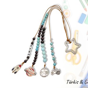 Może przedstawiać: Zestaw czterech kolorowych naszyjników z koralików z zawieszkami, w tym rakietą, planetą, piłką nożną i traktorem. Naszyjniki są wykonane z turkusowych i brązowych koralików i mają srebrny zawieszkę w kształcie gwiazdy na górze. Tekst "NAME" jest napisany na jednym z naszyjników.