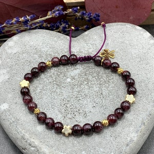 Könnte beinhalten: Ein Armband aus roten Edelsteinen mit goldenen Blumenanhängern und einem goldenen Kleeblatt-Anhänger. Das Armband ist aus einem violetten Band gefertigt und hat einen verschiebbaren Knotenverschluss.