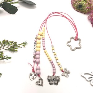 Może przedstawiać: Naszyjnik z różowych i żółtych koralików z srebrnym zawieszką w kształcie motyla, serca i kwiatu. Naszyjnik ma również napis "NAME" wyłożony białymi koralikami.