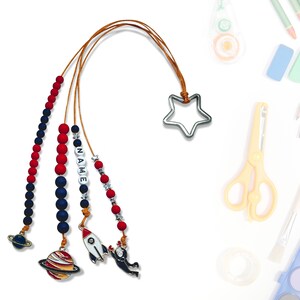 Może przedstawiać: Kolorowa zakładka z koralików z zawieszkami w kształcie gwiazdy, rakiety, astronauty i planety. Koraliki są czerwone, niebieskie i białe. Zakładka ma również napis "NAME" wyłożony białymi koralikami.